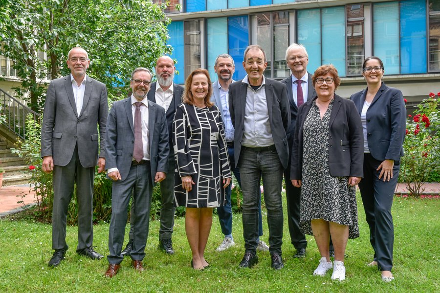 2 Frauen und 7 Männer stehen in einer Gruppe auf einer grünen Wiese vor einem Gebäude. Sie blicken lächelnd in die Kamera.  2 Frauen und 7 Männer stehen in einer Gruppe auf einer grünen Wiese vor einem Gebäude. Sie blicken lächelnd in die Kamera.