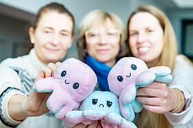 Drei Frauen halten lächelnde Stoff-Kraken in die Kamera; die Figuren sind rosa und blau und dienen als Maskottchen eines Angebots der Psychiatrischen Institutsambulanz für Kinder.