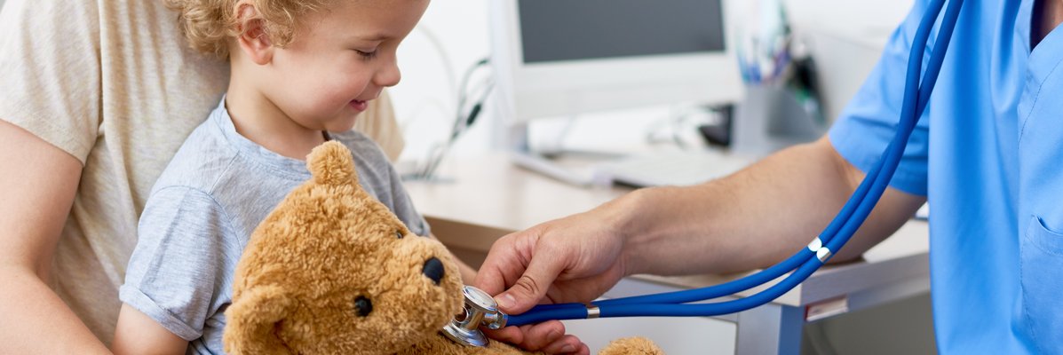 Mann in blauem Kittel hält ein Stetoskop an einen Teddy, den eine Kind auf dem Schoß einer Frau hält