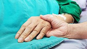 Eine Hand hält die Hand einer Person, die offenbar in einem Krankenbett liegt und eine Flexüle im Arm hat.