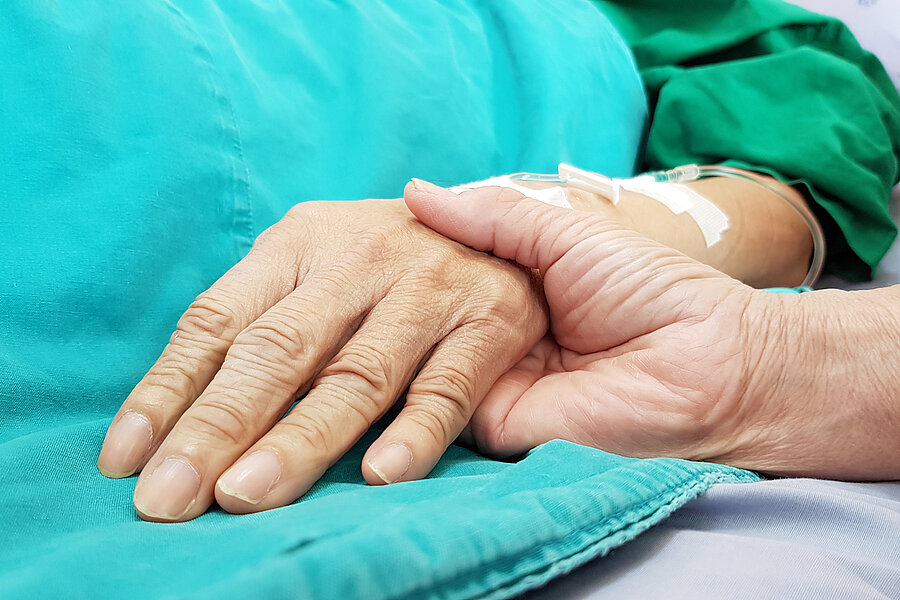 Eine Hand hält die Hand einer Person, die offenbar in einem Krankenbett liegt und eine Flexüle im Arm hat. Eine Hand hält die Hand einer Person, die offenbar in einem Krankenbett liegt und eine Flexüle im Arm hat.