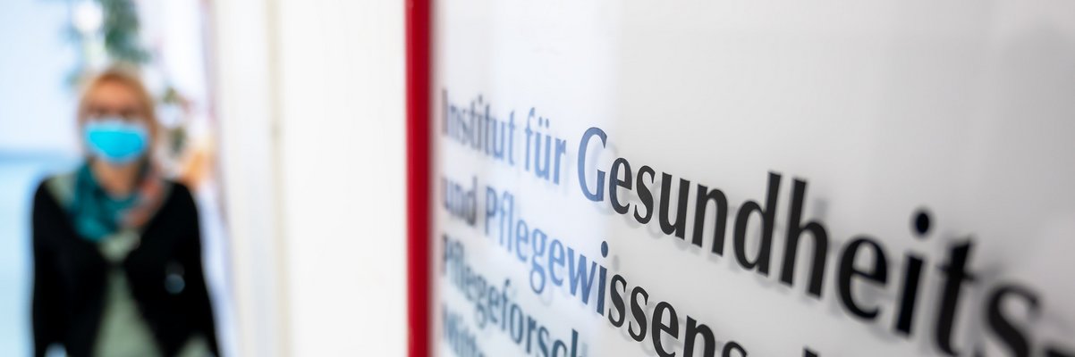 Schild mit Aufschrift Institut für Gesundheits- und Pflegewissenschaft. Eine Frau kommt den Gang entlang