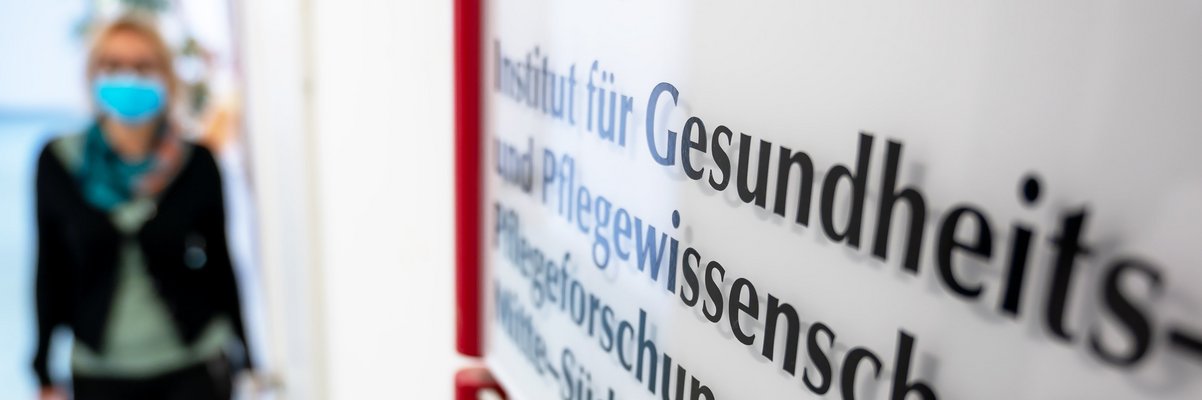 Schild mit Aufschrift Institut für Gesundheits- und Pflegewissenschaft. Eine Frau kommt den Gang entlang