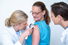 Eine Person links im Bild untersucht den Oberarm einer anderen Person, die in der Mitte des Bildes steht, mit einem Dermatoskop. Eine weitere Person steht rechts im Bild und schaut zu. Alle tragen medizinische Berufskleidung.