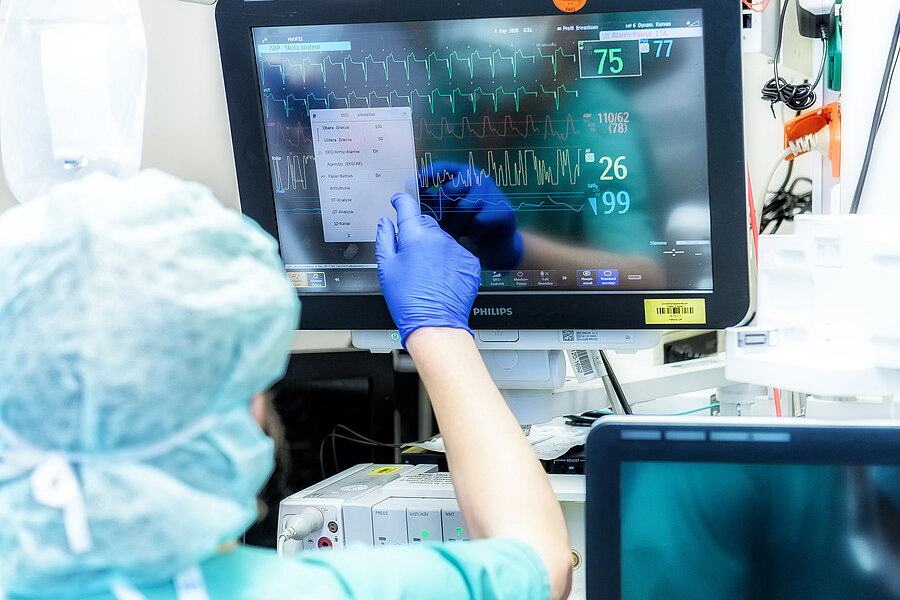 Eine Person, die eine OB-Haube und medizinische Einmalhandschuhe trägt, bedient einen Bildschirm. Auf dem Display sind verschiedene Vitalparameter wie Puls, Blutdruck und Sauerstoffsättigung erkennbar.  Eine Person, die eine OB-Haube und medizinische Einmalhandschuhe trägt, bedient einen Bildschirm. Auf dem Display sind verschiedene Vitalparameter wie Puls, Blutdruck und Sauerstoffsättigung erkennbar.