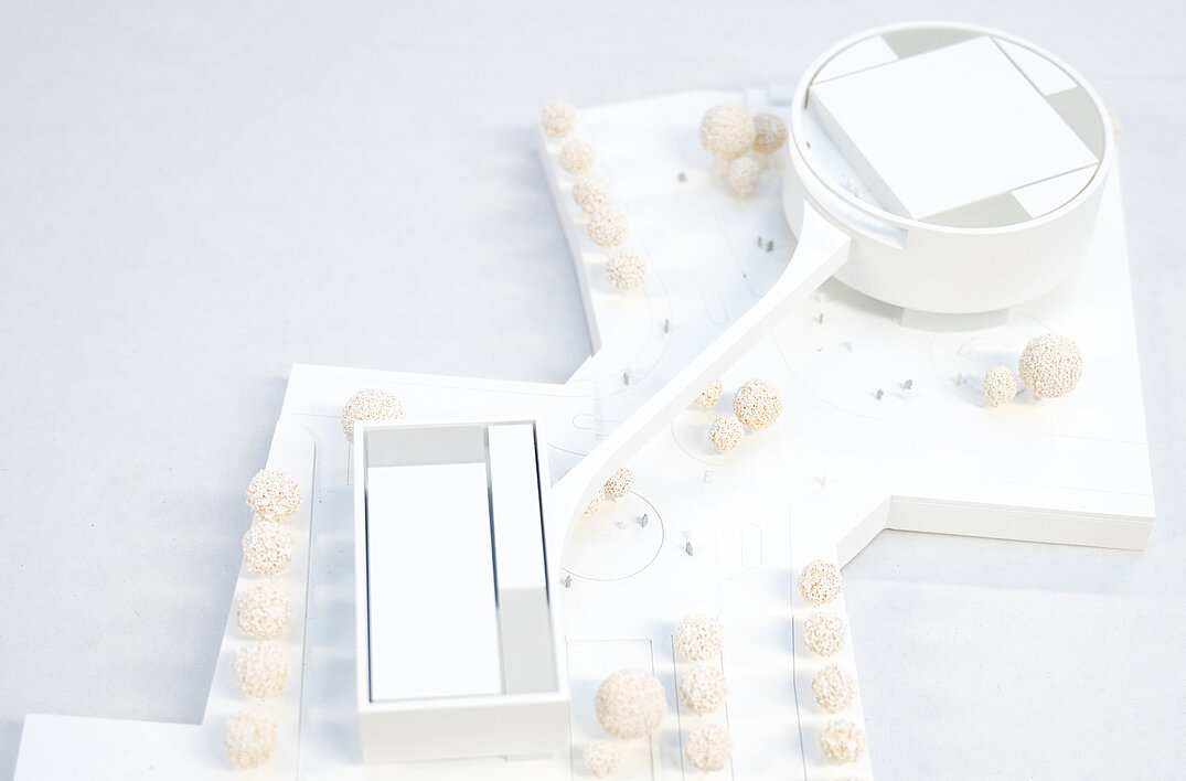 Detailaufnahme des 3-D-Modells des PRZ-Komplexes in weiß. Zusehen sind von schräg oben das rechteckige PRZ 1 und das runde PRZ 1, verbunden mit der Fußgängerbrücke und umgeben von einfarbigen Holzbäumchen.