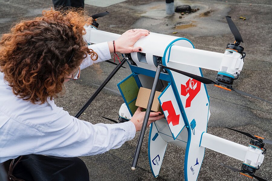 Eine Frau verstaut einen kleinen Pappkarton in einer Drohne. Die Drohne ist etwas größer als ein Umzugskarton, hat acht handgroße Rotoren und ist auf der Seite mit einem knallroten Apothekensymbol versehen.  Eine Frau verstaut einen kleinen Pappkarton in einer Drohne. Die Drohne ist etwas größer als ein Umzugskarton, hat acht handgroße Rotoren und ist auf der Seite mit einem knallroten Apothekensymbol versehen.
