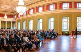 Mehrere Hundert Menschen in der Aula des Löwengebäudes, während eine Frau am Podium spricht.