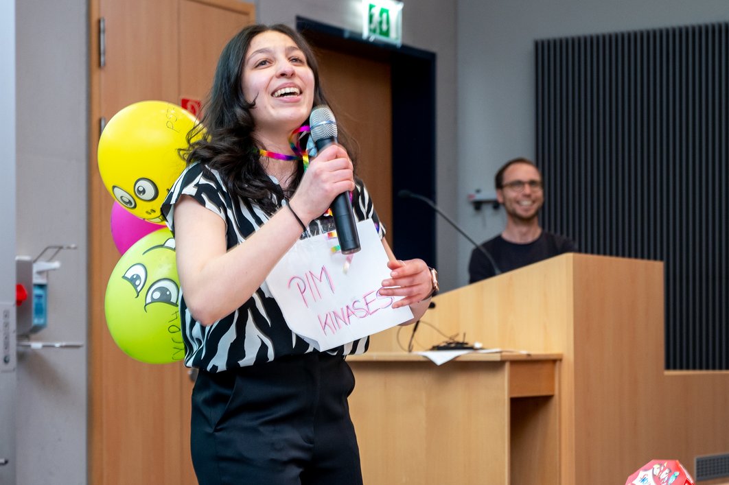 Eine junge Person hält ein Mikro in der Hand. Auf ihren Rücken sind mehrere bunte Luftballon befestigt. Vor ihrem Köper hängt ein Schild mit der Aufschrift "PIM KINASES", die Teil der Erzählung waren. 