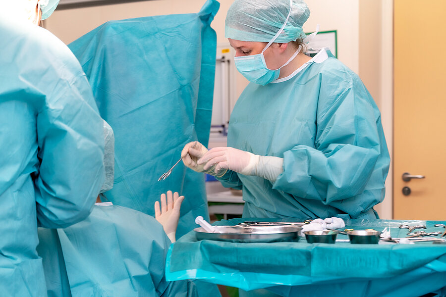 Eine Person in OP-Kleidung steht an einem OP-Tisch und hält ein Instrument in der Hand. Auf dem Tisch vor ihr liegen Instrumente für die OP. Links im Bild befinden sich im Anschnitt zwei weitere Personen in OP-Kleidung. Eine Person in OP-Kleidung steht an einem OP-Tisch und hält ein Instrument in der Hand. Auf dem Tisch vor ihr liegen Instrumente für die OP. Links im Bild befinden sich im Anschnitt zwei weitere Personen in OP-Kleidung.