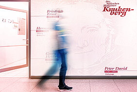 Eine Person läuft einen Flur entlang. Die Person befindet sich in der Bewegung und ist nur schemenhaft dargestellt. Im Hintergrund befindet sich eine Wand mit dem Schriftzug "Die halleschen Mediziner Krukenberg" sowie weiteren Namen und Daten.