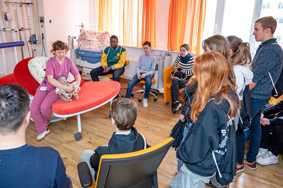 Eine Gruppe von Schüler:innen steht im Kreißsaal des Universitätsklinikums Halle (Saale). Hebamme Astrid Dening erklärt den Schüler:innen die Abläufe und den Geburtsvorgang.  Eine Gruppe von Schüler:innen steht im Kreißsaal des Universitätsklinikums Halle (Saale). Hebamme Astrid Dening erklärt den Schüler:innen die Abläufe und den Geburtsvorgang.