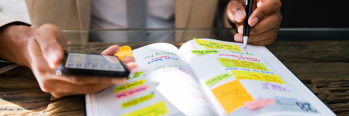 Ein aufgeschlagener Kalender mit vielen Termin. Ein Mensch hält einen Stift und ein Handy und schaut sich die Termine an