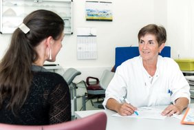 Frau in weißem Arztkittel an einem Schreibtisch. Ihr gegen über eine Frau mit langen dunklen Haaren, die nur von hinten zu sehen ist