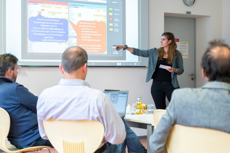 Eine weibliche Person referiert im Rahmen der Kickoff-Veranstaltung zum PEAK-Projekt zu einem Sachverhalt, der an die Wand projiziert ist. Im Vordergrund sind drei Personen von hinten zu sehen, die ihr zuhören.   Eine weibliche Person referiert im Rahmen der Kickoff-Veranstaltung zum PEAK-Projekt zu einem Sachverhalt, der an die Wand projiziert ist. Im Vordergrund sind drei Personen von hinten zu sehen, die ihr zuhören. 