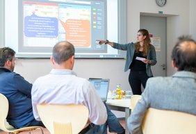 Eine weibliche Person referiert im Rahmen der Kickoff-Veranstaltung zum PEAK-Projekt zu einem Sachverhalt, der an die Wand projiziert ist. Im Vordergrund sind drei Personen von hinten zu sehen, die ihr zuhören. 