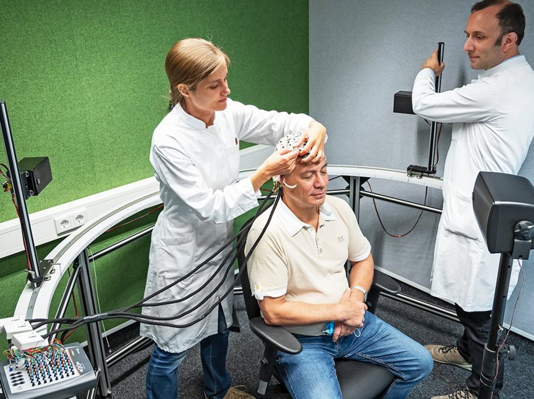 Eine Frau setzt einem Patienten eine Haube mit Leitungen auf en Kopf. ein Arzt stellt die Geräte für eine Messung ein