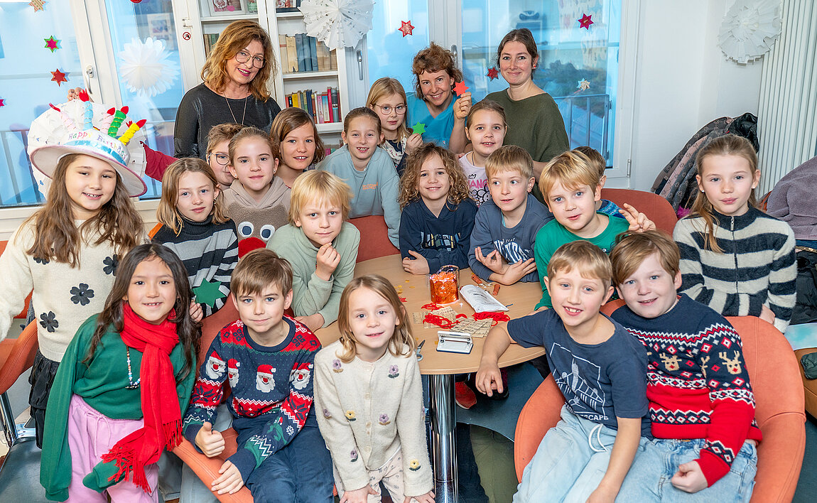 Gruppenfoto einer Schulklasse. Die Kinder befinden sich in einem weihnachtlich geschmückten Aufenthaltsraum auf einer Station eines Krankenhauses. Im Hintergrund stehen drei erwachsene Personen.
