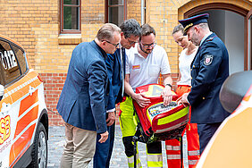 Fünf Personen – zwei in Rettungsuniform, eine in Feuerwehruniform, eine im Anzug und eine in Freizeitkleidung – betrachten gemeinsam einen geöffneten roten Notfallrucksack. Daneben steht ein Einsatzfahrzeug mit der Notrufnummer 112.