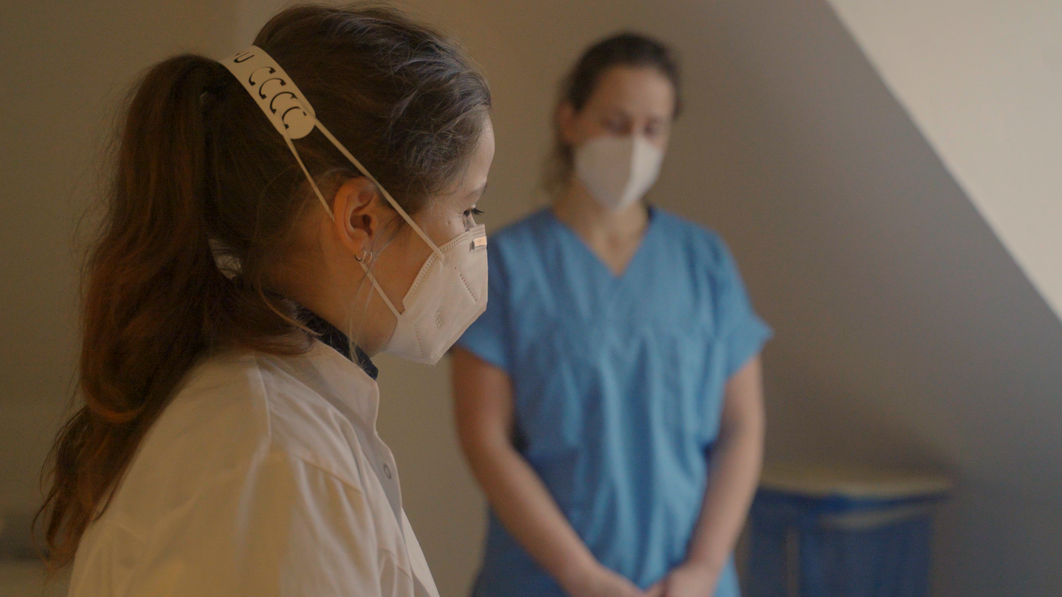 Zwei Frauen in medizinischer Arbeitskleidung stehen in einem Raum, eine im Vordergrund und eine unscharf im Hintergrund.