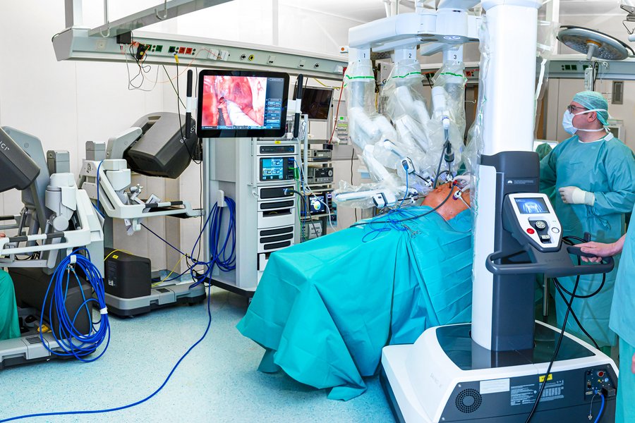 Blick in einen OP-Saal mit dem da Vinci-Roboter, dessen Arme über einem Patienten schweben. Man sieht Chirurgen, die an Konsolen den Roboter bedienen, ein Chirurg steht neben dem Patienten. Vom Patienten ist nur der Bauch zu sehen, der für die Operation aufgebläht ist. Auf einem Monitor ist das Innere des Bauchraums zu sehen.  Blick in einen OP-Saal mit dem da Vinci-Roboter, dessen Arme über einem Patienten schweben. Man sieht Chirurgen, die an Konsolen den Roboter bedienen, ein Chirurg steht neben dem Patienten. Vom Patienten ist nur der Bauch zu sehen, der für die Operation aufgebläht ist. Auf einem Monitor ist das Innere des Bauchraums zu sehen.