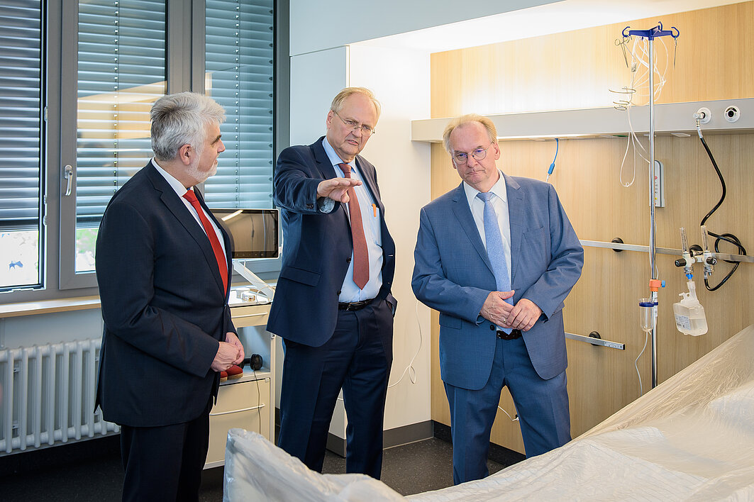 Prof. Dr. Thomas Moesta steht zwischen Prof. Dr. Armin Willingmann und Reiner Haseloff in einem Patient:innenzimmer und gestikuliert.