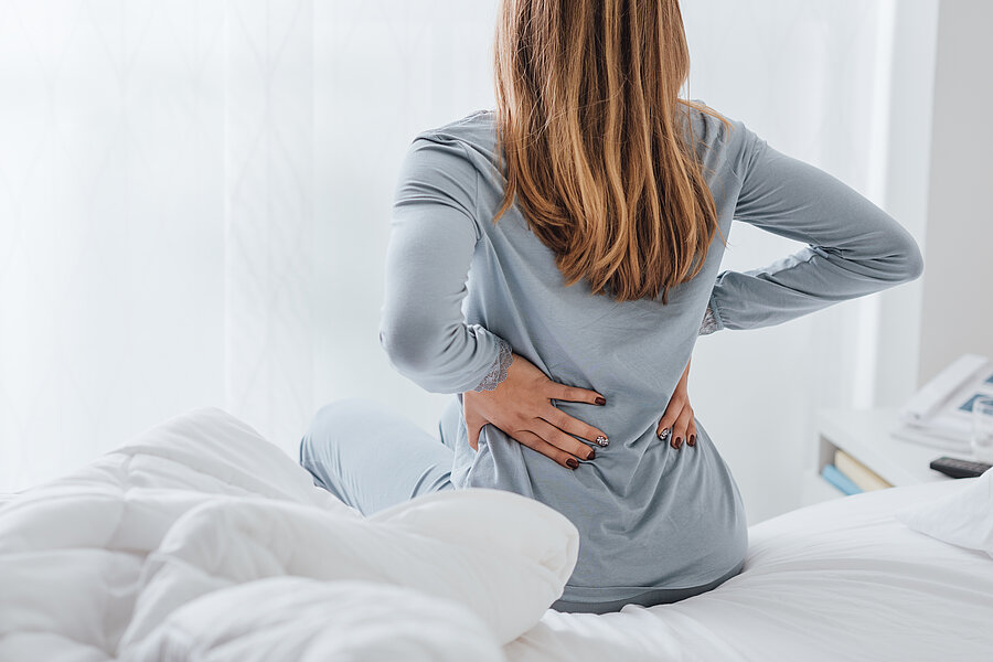 Frau von hinten auf einem Bett sitzend. Sie fasst sich mit beiden Henden an den unteren Rücken.  Frau von hinten auf einem Bett sitzend. Sie fasst sich mit beiden Henden an den unteren Rücken.