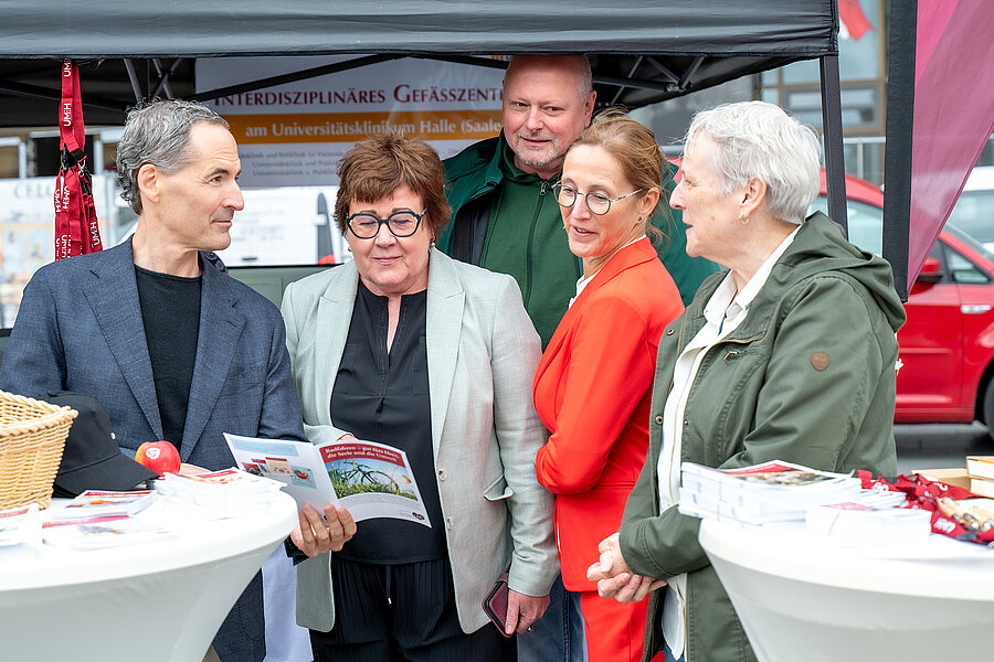 Fünf Personen stehen an einem Infostand an zwei Stehtischen. Eine Person hält einen Flyer in der Hand.  Fünf Personen stehen an einem Infostand an zwei Stehtischen. Eine Person hält einen Flyer in der Hand.