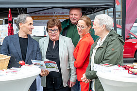 Fünf Personen stehen an einem Infostand an zwei Stehtischen. Eine Person hält einen Flyer in der Hand.