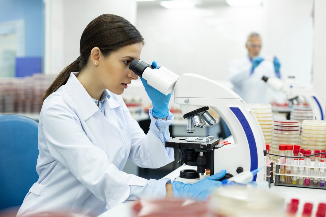 Frau im weißen Kittel sitzt an einem Mikroskop im Labor, im Hintergrund arbeitet ein Mitarbeiter
