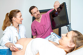 Eine Ärztin untersucht den Bauch einer schwangeren Frau mithilfe eines Ultraschallgeräts. Die schwangere Frau liegt auf einer Liege. Die Ärztin und ein weiterer Arzt schauen sich die Ultraschallbilder auf einem Monitor an.