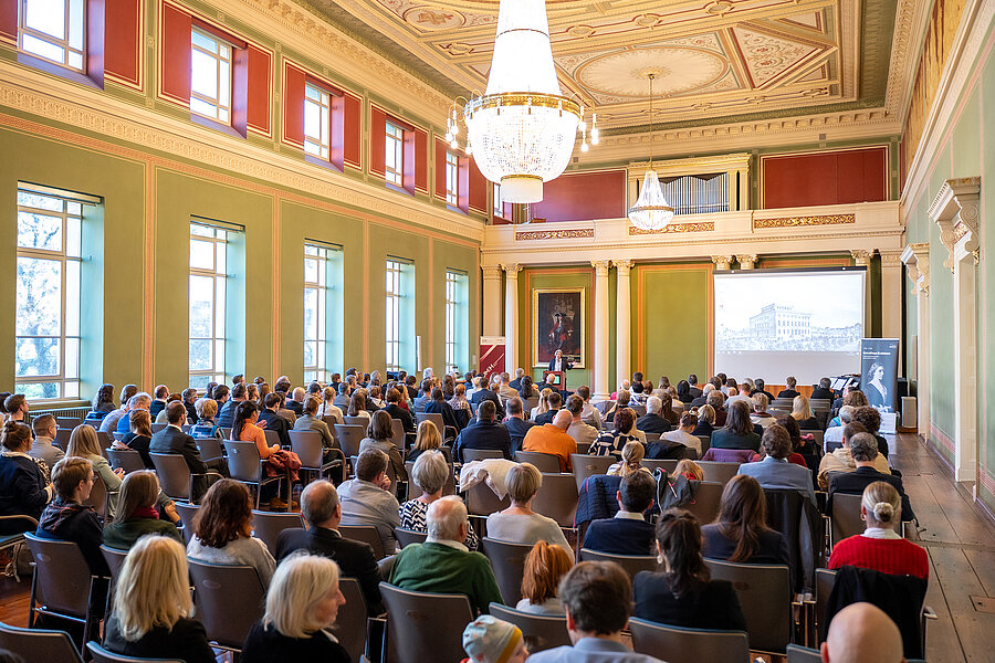 Ein Blick in den gefüllten Festsaal des Löwengebäudes am Uniplatz der Martin-Luther-Universität Halle-Wittenberg. Am weit entfernten Pult ist eine Frau zu erkennen, die spricht und dabei gestikuliert. Neben ihr wird ein historisches Bild des Löwengebäudes präsentiert.  Ein Blick in den gefüllten Festsaal des Löwengebäudes am Uniplatz der Martin-Luther-Universität Halle-Wittenberg. Am weit entfernten Pult ist eine Frau zu erkennen, die spricht und dabei gestikuliert. Neben ihr wird ein historisches Bild des Löwengebäudes präsentiert.