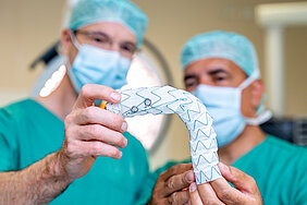 Zwei Personen in OP-Kleidung halten einen Aorten-Stent in die Kamera. Hierbei handelt es sich um ein längliches bewegliches Röhrchen. Auf dem Röhrchen sind Auslassungen angezeichnet.