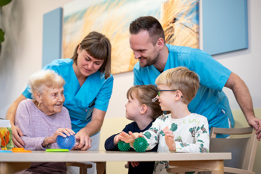 Zwei Kinder und eine ältere Dame sitzen an einem Tisch und schauen sich an. Alle halten Massagebälle in den Händen. Hinter ihnen stehen zwei Personen in medizinischer Berufskleidung und interagieren mit ihnen.  Zwei Kinder und eine ältere Dame sitzen an einem Tisch und schauen sich an. Alle halten Massagebälle in den Händen. Hinter ihnen stehen zwei Personen in medizinischer Berufskleidung und interagieren mit ihnen.