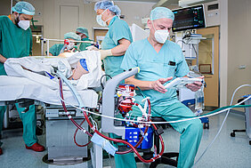 Vier Personen in medizinischer Berufskleidung versorgen eine Person auf einer Intensivstation in einem Krankenhaus. Im Vordergrund befindet sich ein ECMO-System. Dahinter sitzt eine weitere Person in medizinischer Berufskleidung auf einem Hocker und hält ein Tablet in der Hand.