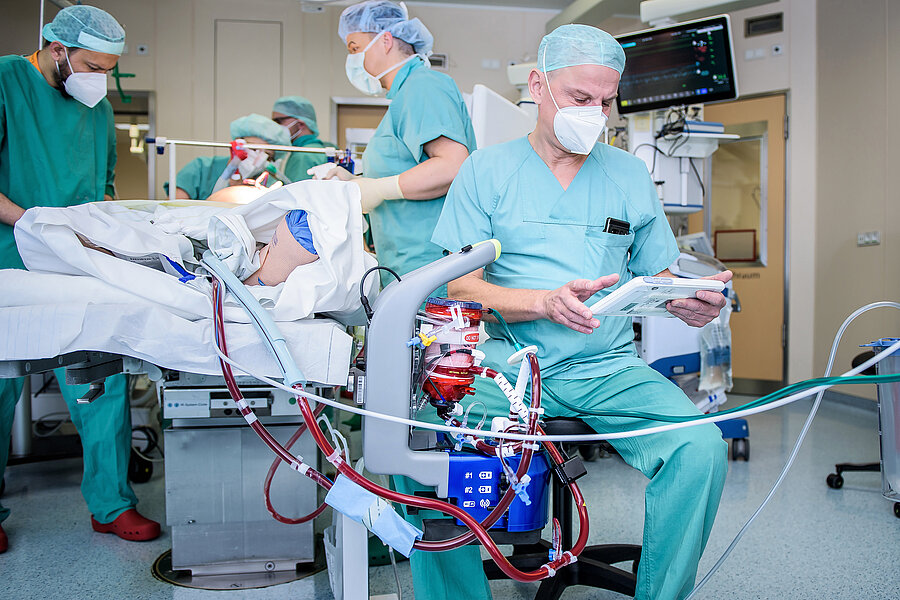 Vier Personen in medizinischer Berufskleidung versorgen eine Person auf einer Intensivstation in einem Krankenhaus. Im Vordergrund befindet sich ein ECMO-System. Dahinter sitzt eine weitere Person in medizinischer Berufskleidung auf einem Hocker und hält ein Tablet in der Hand.  Vier Personen in medizinischer Berufskleidung versorgen eine Person auf einer Intensivstation in einem Krankenhaus. Im Vordergrund befindet sich ein ECMO-System. Dahinter sitzt eine weitere Person in medizinischer Berufskleidung auf einem Hocker und hält ein Tablet in der Hand.