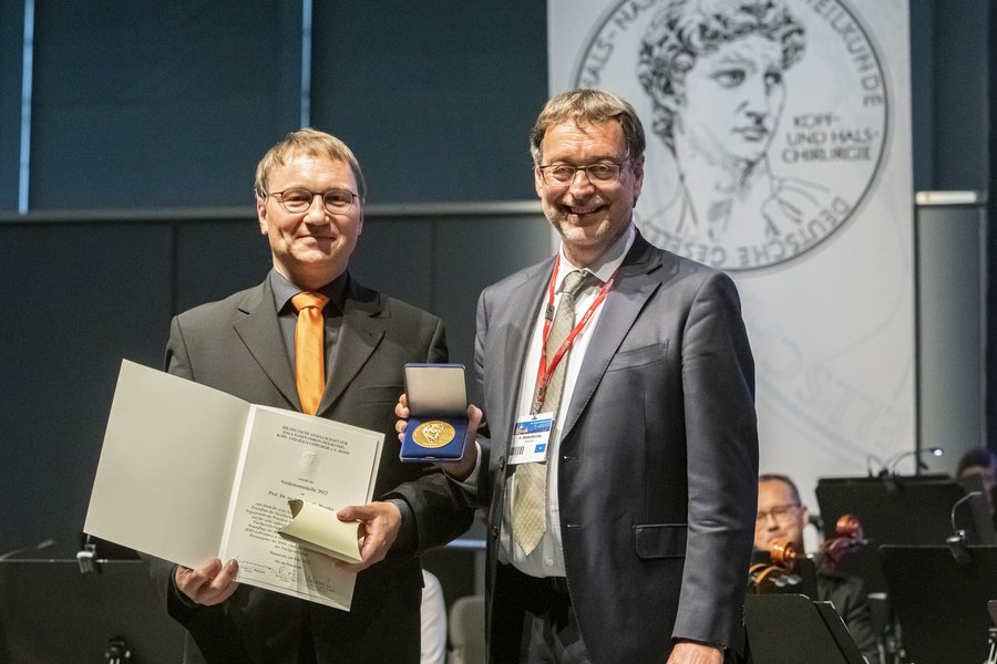 Zwei Herren im Anzug halten eine Urkunde und eine Medaille lächelnd in die Kamera  Zwei Herren im Anzug halten eine Urkunde und eine Medaille lächelnd in die Kamera