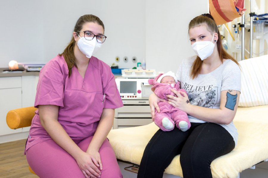 Eine Mutter sitzt mit ihrem Neugeborenen auf einer Liege. Daneben eine Hebamme.  Eine Mutter sitzt mit ihrem Neugeborenen auf einer Liege. Daneben eine Hebamme.