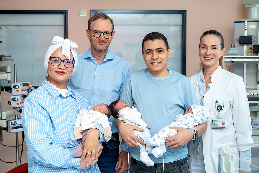 Ein Arzt, eine Ärztin und ein Elternpaar stehen in einem Klinikzimmer. Die Mutter hält ein Baby auf dem Arm, der Vater hält zwei weitere Babys in den Armen. Ein Arzt, eine Ärztin und ein Elternpaar stehen in einem Klinikzimmer. Die Mutter hält ein Baby auf dem Arm, der Vater hält zwei weitere Babys in den Armen.