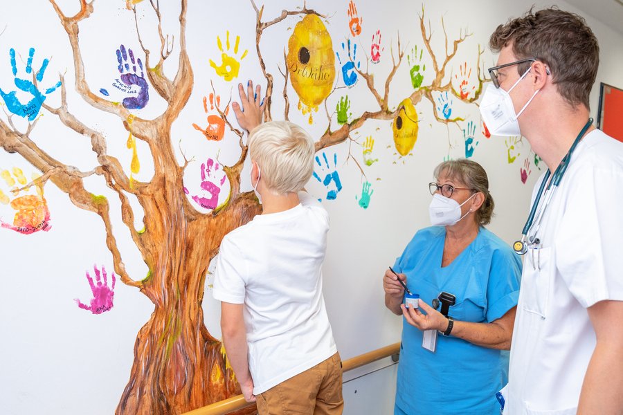 Ein junge mit blonden Haaren und weißem T-Shirt interlässt einen blauen Handabdruck an einem gemalten Baum an der Wand. Neben ihm steht ein Arzt und eine Pflegefachfrau.  Ein junge mit blonden Haaren und weißem T-Shirt interlässt einen blauen Handabdruck an einem gemalten Baum an der Wand. Neben ihm steht ein Arzt und eine Pflegefachfrau.