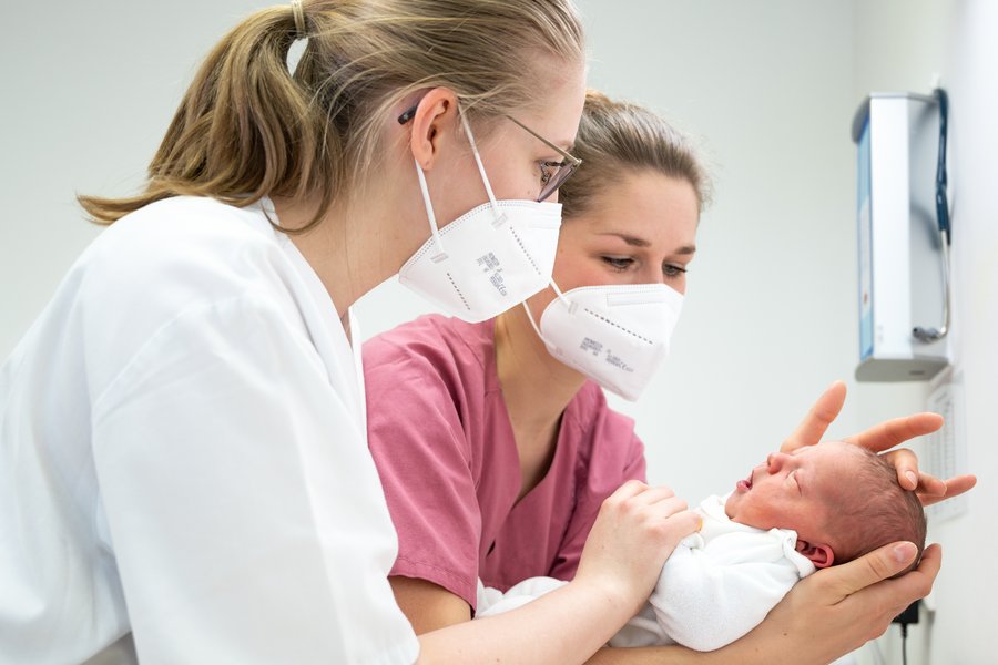 zwei Frauen in weißer und brombeerfarbener medizinischer Arbeitskleidung halten ein neugeborenes Baby  zwei Frauen in weißer und brombeerfarbener medizinischer Arbeitskleidung halten ein neugeborenes Baby