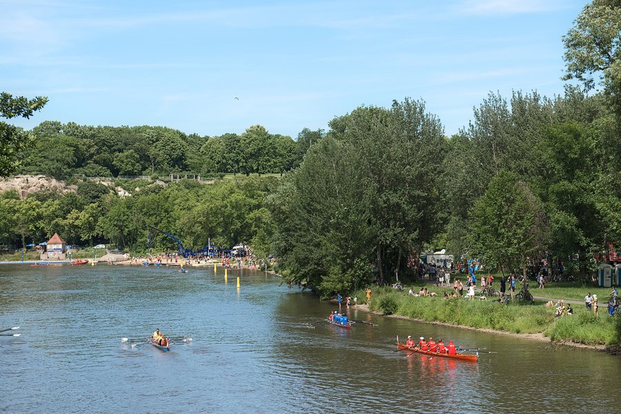 Abschnitt der Saale. Am Ufer sind Ruderboote und Menschen zu erkennen. Im Vordergrund rahmen Bäume das Foto ein.  Abschnitt der Saale. Am Ufer sind Ruderboote und Menschen zu erkennen. Im Vordergrund rahmen Bäume das Foto ein.