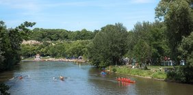 Abschnitt der Saale. Am Ufer sind Ruderboote und Menschen zu erkennen. Im Vordergrund rahmen Bäume das Foto ein.