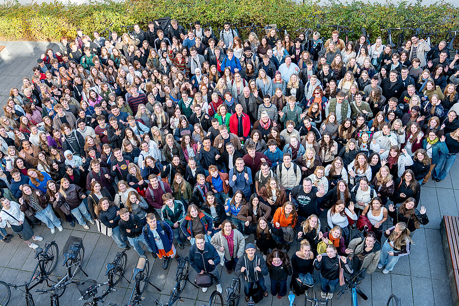 In der Mitte eines Innenhofs stehen mehr als 300 Personen und blicken nach oben zu einer höher gelegenen Kamera. Einige heben die Hände, andere lächeln fröhlich. In der Mitte eines Innenhofs stehen mehr als 300 Personen und blicken nach oben zu einer höher gelegenen Kamera. Einige heben die Hände, andere lächeln fröhlich.