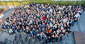 In der Mitte eines Innenhofs stehen mehr als 300 Personen und blicken nach oben zu einer höher gelegenen Kamera. Einige heben die Hände, andere lächeln fröhlich.