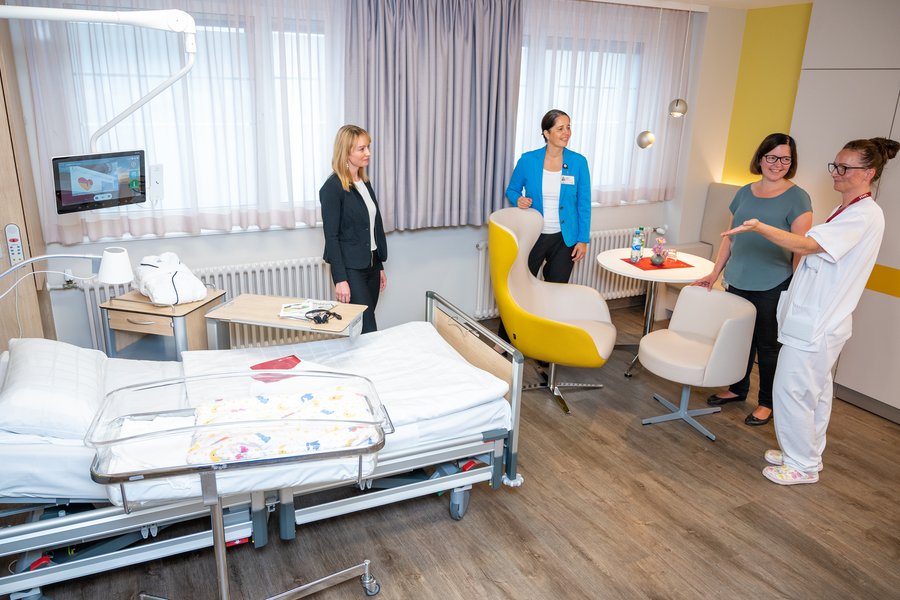 Krankenhauszimmer von schräg oben. Links ist ein Krankenbett zu sehen. im rechten oberen Bildrand stehen vier Frauen an einer gelben Sitzgruppe in einem Gespräch zusammen.  Krankenhauszimmer von schräg oben. Links ist ein Krankenbett zu sehen. im rechten oberen Bildrand stehen vier Frauen an einer gelben Sitzgruppe in einem Gespräch zusammen.