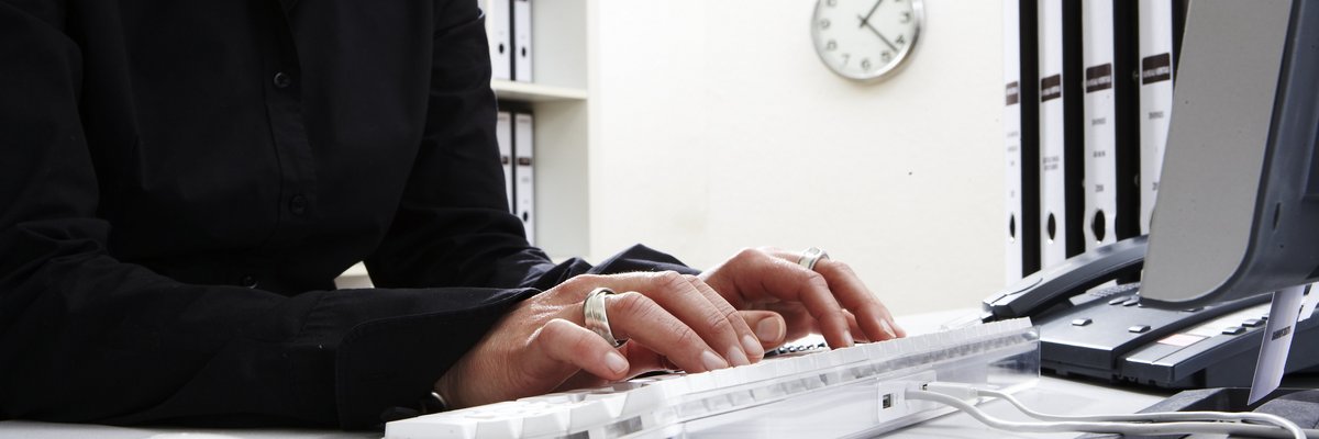 Frau schreibt auf einer Computertastatur am Schreibtisch, auf dem Aktenordner stehen. Im Hitnergrund ein Regal mit Ordnern