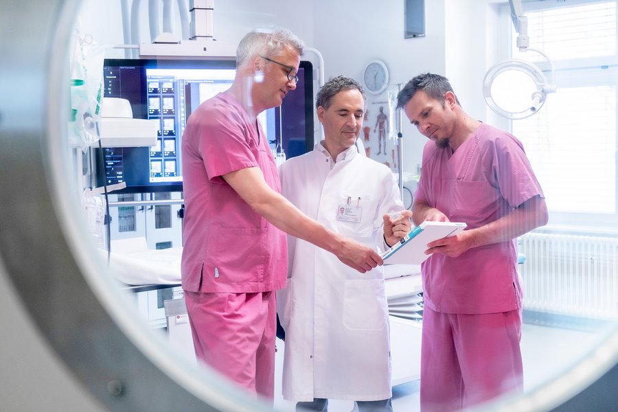 Durch ein Bullauge geht der Blick in ein Herzkatheterlabor. Darin stehen drei Männer in medizinsicher Arbeitskleidung. Sie blicken gemeinsam auf Unterlagen und den Stent.  Durch ein Bullauge geht der Blick in ein Herzkatheterlabor. Darin stehen drei Männer in medizinsicher Arbeitskleidung. Sie blicken gemeinsam auf Unterlagen und den Stent.