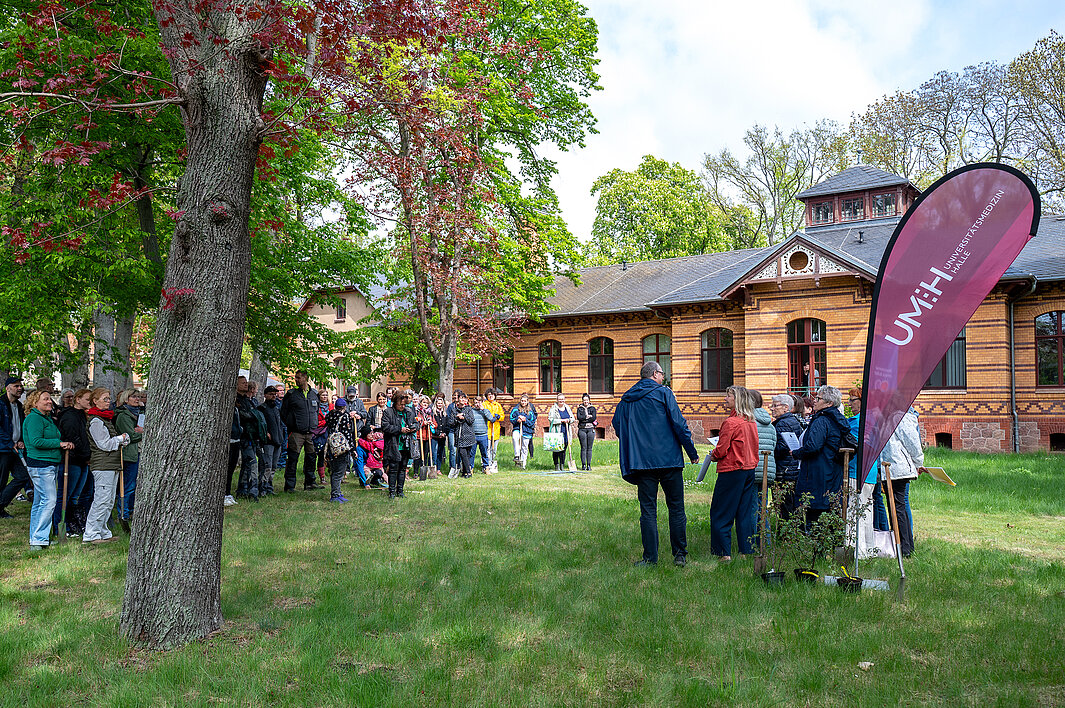 Viele Personen stehen im Garten der Psychiatrischen Klinik der Universitätsmedizin Halle. Rechts stehen ein Chor und der Klinikdirektor. Ihnen gegenüber stehen Teilnehmende der Pflanzaktion. Links im Bild befindet sich ein großer Baum, im Hintergrund ein Teil des Klinikgebäudes.