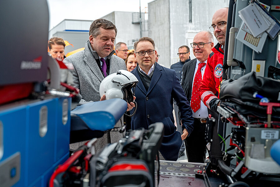 Mehrere Personen schauen in das Innere eines Rettungshubschraubers. Die Person links im Bild hält einen Helm in den Händen.  Mehrere Personen schauen in das Innere eines Rettungshubschraubers. Die Person links im Bild hält einen Helm in den Händen.